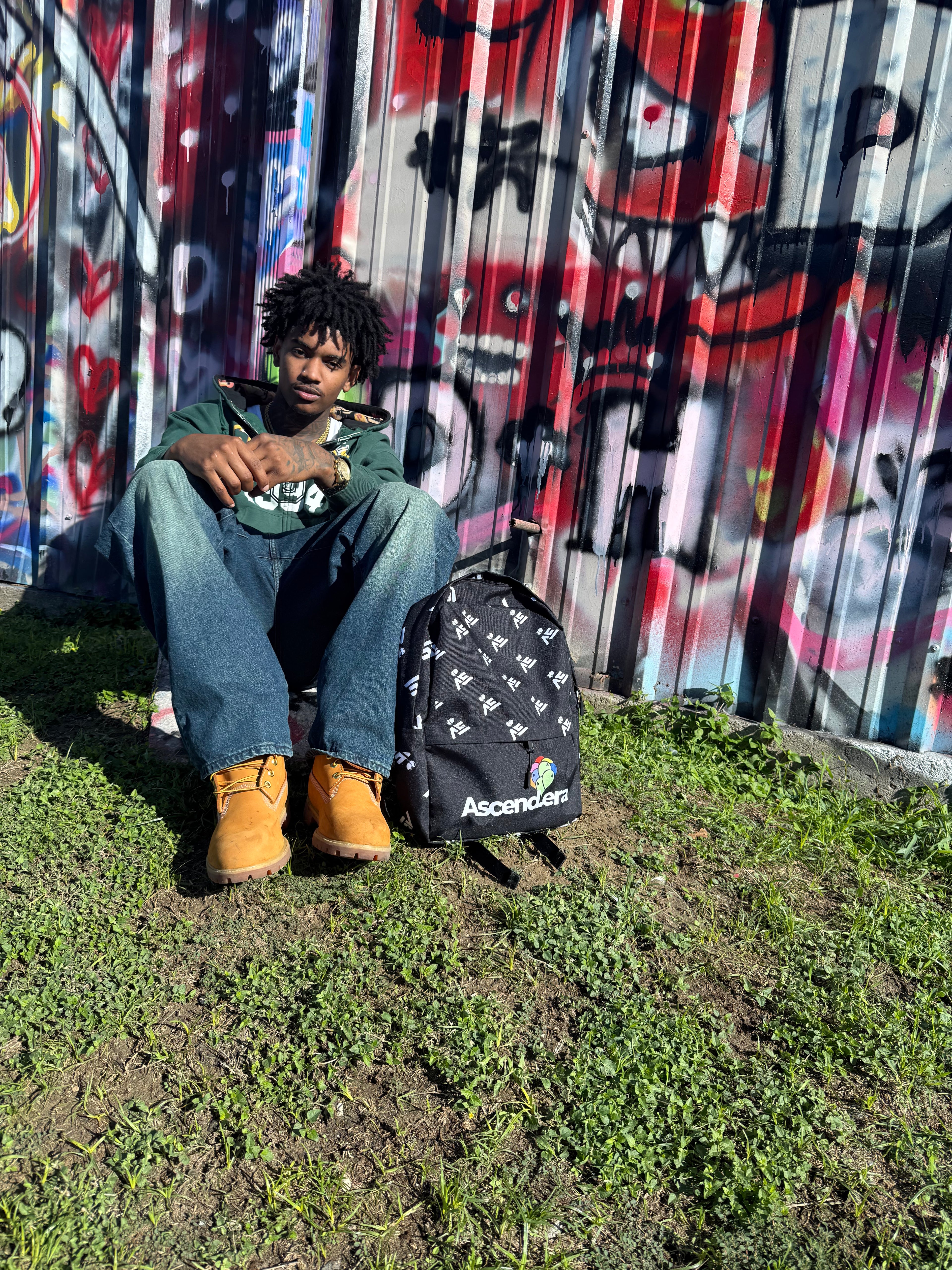 Person sitting on grass with a backpack in front of a graffiti-covered wall